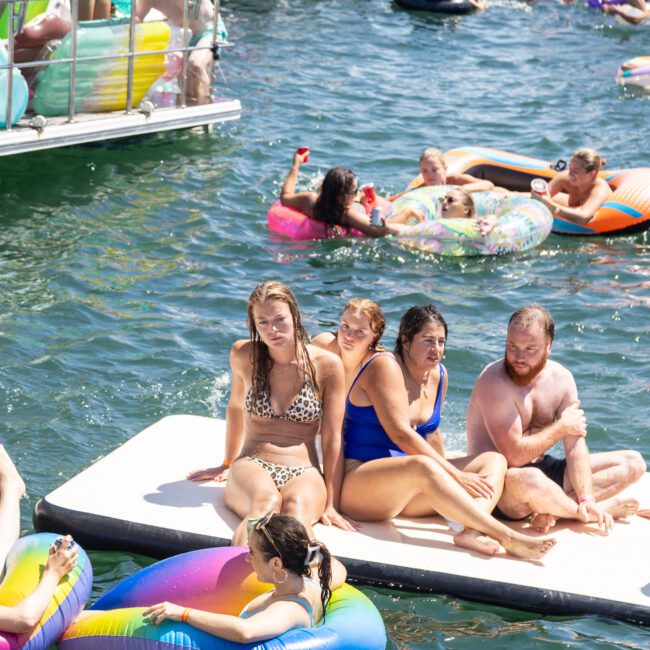 A group of people enjoying a sunny day on inflatable rafts and mats in a lake. Some are sitting and others are floating around in colorful tubes. Boats are nearby, and everyone appears relaxed and cheerful.