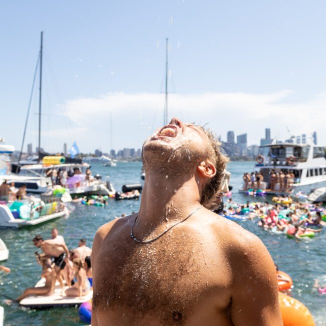 A shirtless man stands on a boat, leaning back with water splashing upwards towards his face. In the background, colorful floats and boats fill a lively waterfront scene under a clear blue sky.