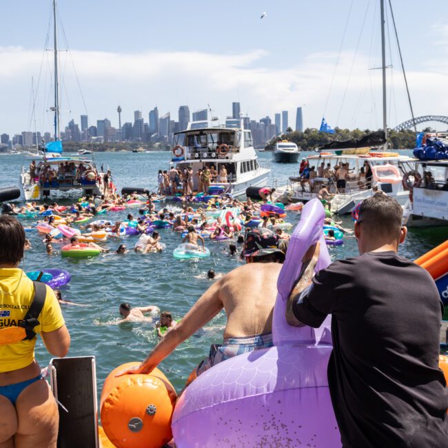 A crowded waterfront scene with people enjoying the water on colorful inflatables. Nearby boats are anchored, and a city skyline with tall buildings is visible in the background. A person is entering the water using a large floating unicorn.