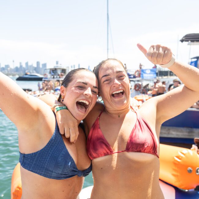 Two smiling women in swimsuits celebrate at a lively beach party with boats and city skyline in the background. One raises her thumb excitedly.