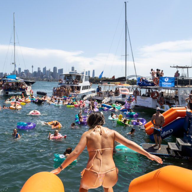 A person in a swimsuit stands on an inflatable slide, overlooking a busy water scene with boats and people on floaties. The city skyline is visible in the background on a sunny day.