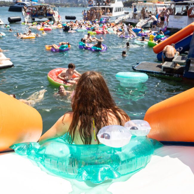 A woman lounges on a giant inflatable, overlooking a lively gathering of people in colorful floats and swimming in a sunlit lake. Boats are visible in the background under a clear blue sky.