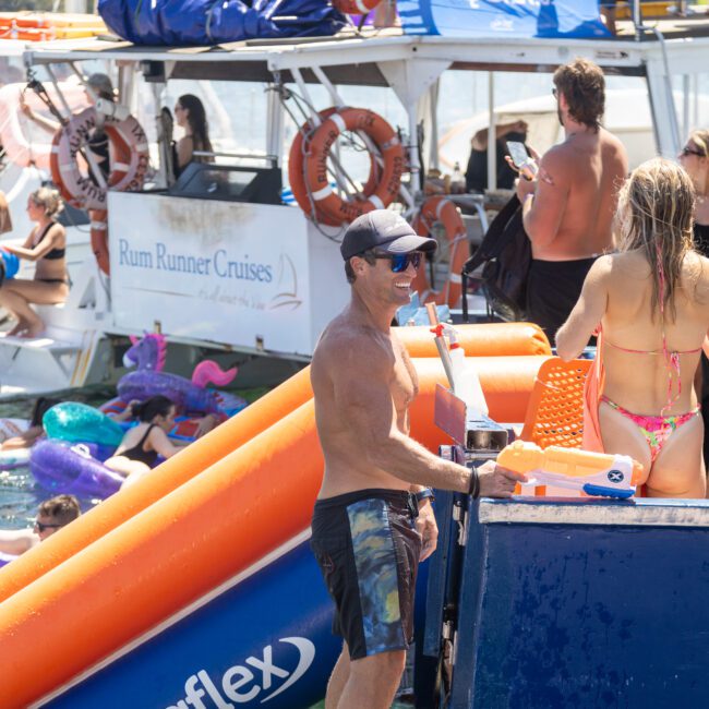 A group of people enjoying a boat party on the water. Some are swimming, while others relax near an inflatable slide on the boat. The boat is equipped with life rings and banners, and a man in swim trunks stands on the deck.