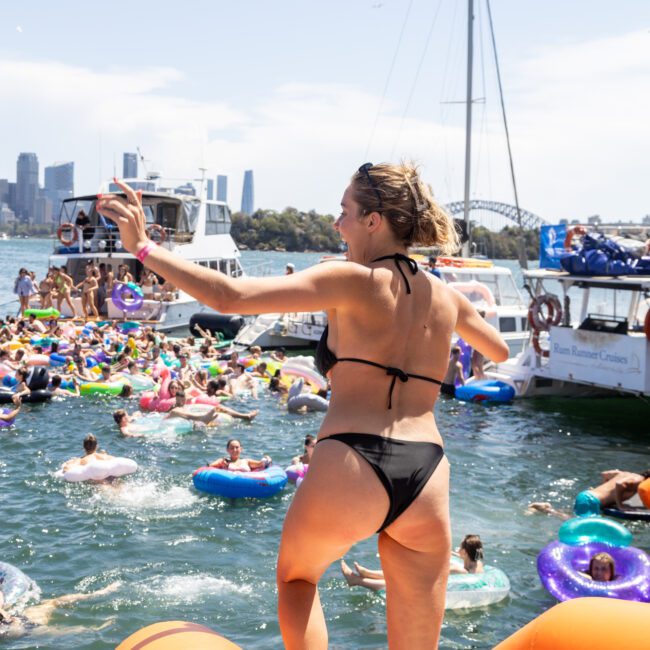 Woman in a black bikini stands on the edge of a yacht, preparing to jump into a crowded group of people in colorful inflatable rings in the water. Other boats and cityscape are visible in the background under a sunny sky.