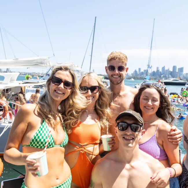 A group of five friends smiling on a boat, wearing swimsuits. They hold drinks and enjoy sunny weather. Sailboats and a city skyline are visible in the background.