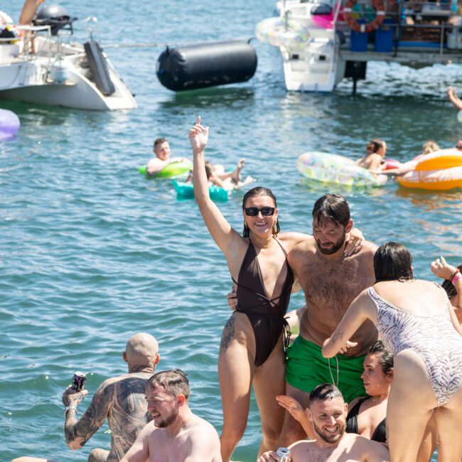 A group of people enjoying a sunny day on the water with boats and inflatables. Some are on a floating platform, smiling and posing. The background shows a city skyline. The atmosphere is lively and festive.