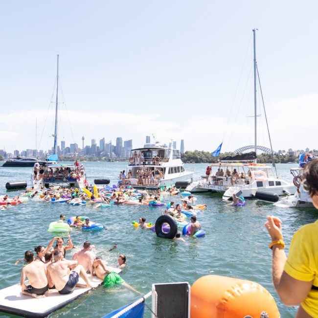 A lively boat party on the water with people enjoying themselves on inflatables and swimming. Boats are anchored nearby, and the city skyline is visible in the background. A lifeguard in a yellow shirt watches the scene.