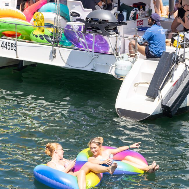 Two people relax on colorful inflatable floats in the water, smiling and pointing, near a boat with several people onboard. The sun is shining brightly, creating a vibrant and cheerful atmosphere.