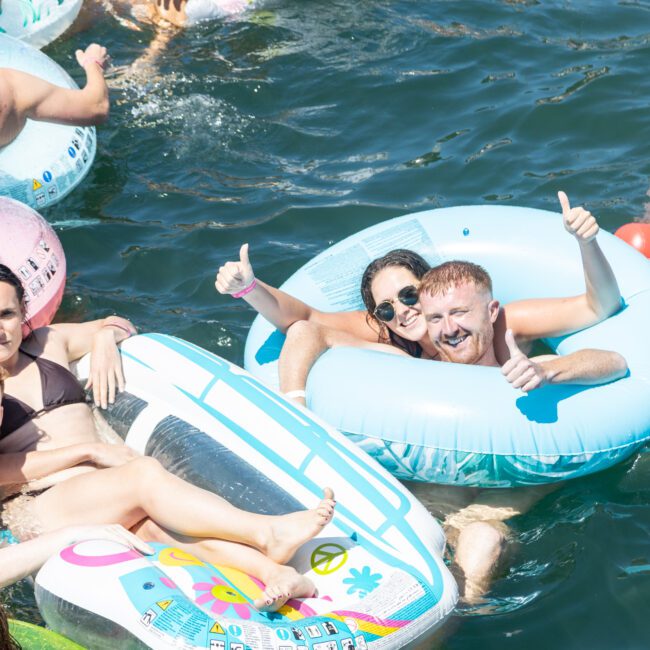 A group of people floating in a lake on inflatable rafts and tubes. Some are giving thumbs up and smiling, enjoying a sunny day in the water. The atmosphere is festive and relaxed.