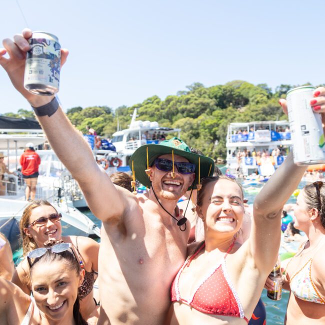 A group of friends in swimsuits smiles and holds up drinks while standing in the water. Boats are visible in the background, with a lively outdoor atmosphere and greenery surrounding the area.