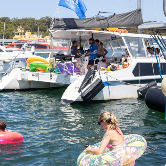 A woman with a floral inflatable ring steps off a sailboat into the water. Others relax onboard the catamaran. A blue flag waves above, and multiple boats are anchored nearby, with a forested shoreline in the background.