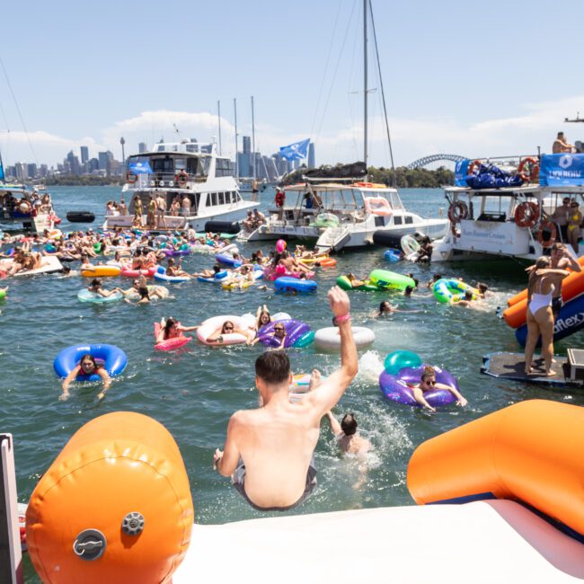 A lively scene of people on colorful inflatables enjoying a sunny day on the water. Boats are anchored nearby, and the city skyline is visible in the background. The atmosphere is festive and relaxed.