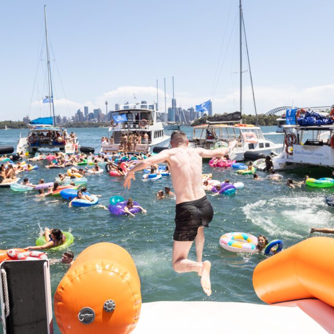 A man jumps off a boat into the water, surrounded by numerous people on colorful inflatable rafts. Several boats are anchored nearby under a clear blue sky, creating a lively party atmosphere on the water.