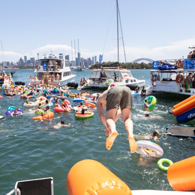 A person dives into a busy water scene from a boat. The water is filled with people on colorful inflatables. Several boats are anchored nearby with a city skyline visible in the background under a bright, sunny sky.