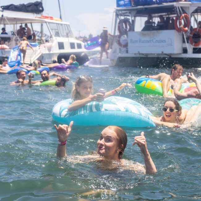 A group of people enjoys a sunny day on the water, floating on colorful inflatables. Boats are in the background, and everyone appears relaxed and happy. One person in the foreground smiles and makes a hand gesture.