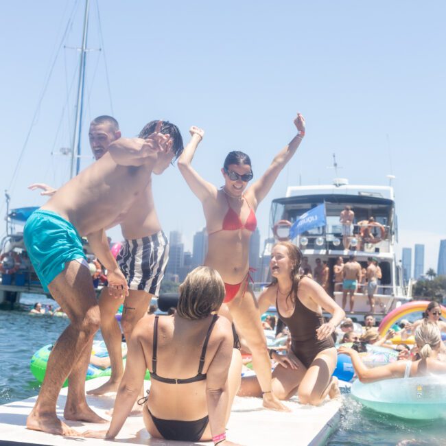 A group of people in swimsuits enjoy a sunny day on a floating platform in the water. Boats and a city skyline are visible in the background. One person is raising their arms in celebration.