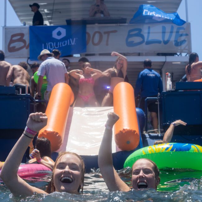 Two people in the water, smiling and raising their arms. In the background, a boat hosts a lively party with people dancing near a slide. Inflatable rings float in the water. A banner on the boat reads "BLOODSHOT BLUE.