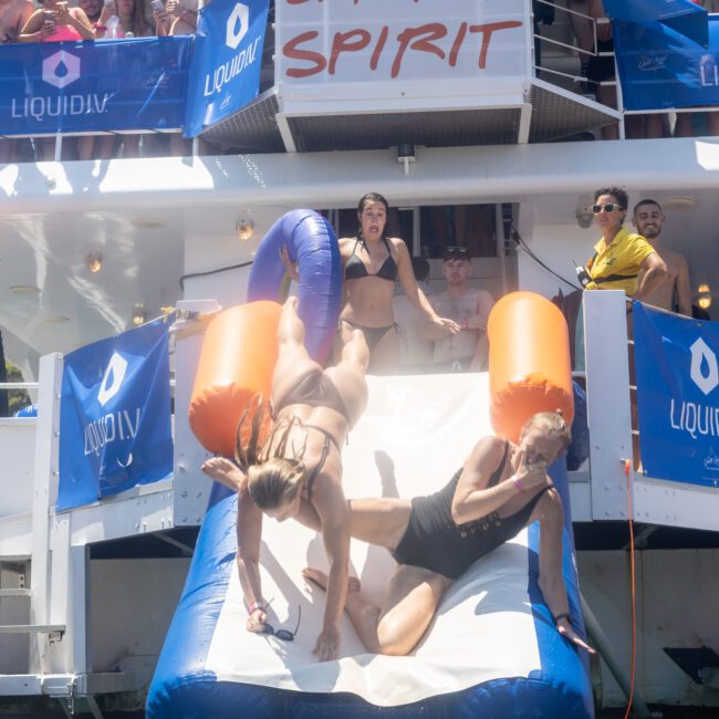 People are sliding down an inflatable slide into the water from a boat with a "Barefoot Spirit" banner. Onlookers watch from the boat's deck, decorated with "LiquidIV" banners. The scene is festive and sunny.