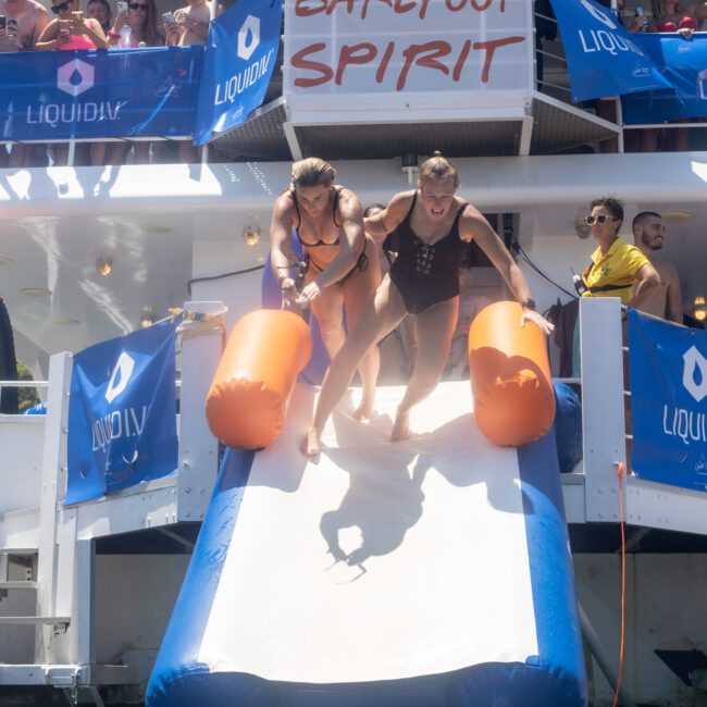 Two people sliding down an inflatable slide off a boat into the water. The boat displays a "Barefoot Spirit" banner, with people watching from above and around. It's a sunny day, and the scene is lively and celebratory.