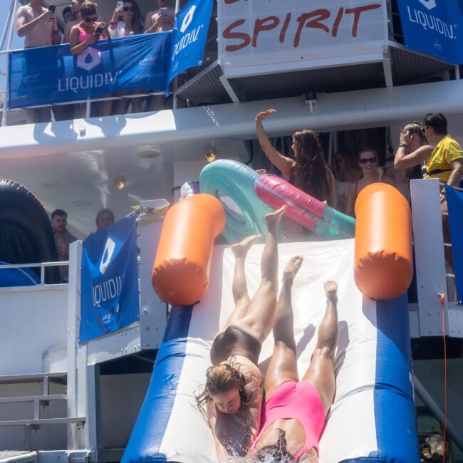 Two people slide down an inflatable water slide from a boat labeled "Barefoot Spirit" into the water. Crowds watch from the boat's deck under banners with "Liquid" logos. The scene is lively and summery.