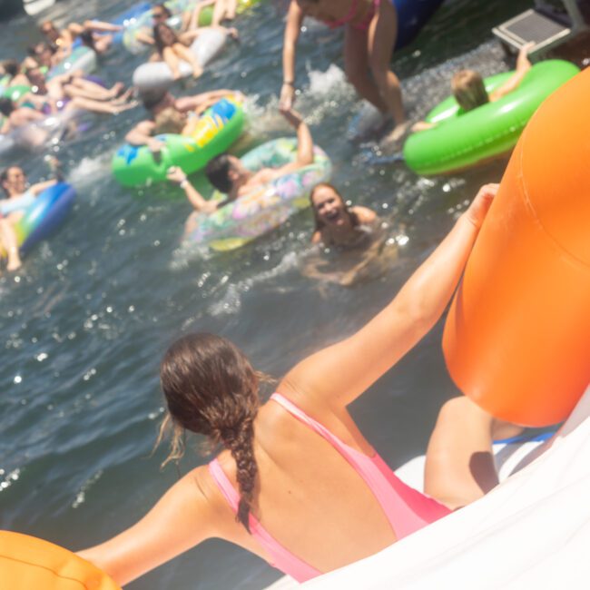 A person in a pink swimsuit sits on an inflatable raft, ready to enter the water. In the background, several people enjoy swimming and relaxing on colorful inflatables in a sunny outdoor setting.