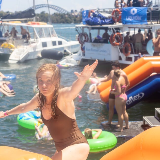 A woman in a brown swimsuit is about to jump into the water from an inflatable obstacle. People are swimming and lounging on floaties in the background, with several boats and a bridge visible in the distance.