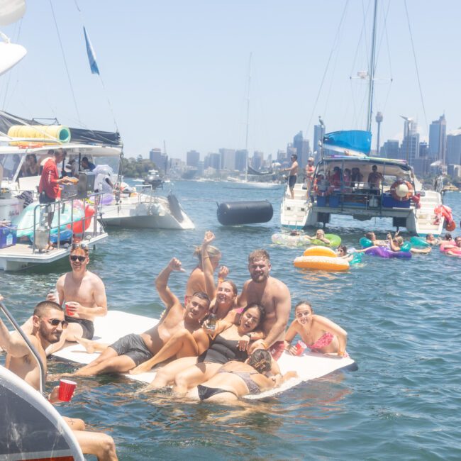 A group of people enjoy a sunny day on the water, lounging on floating mats near boats. The city skyline is visible in the background. Some hold drinks, and inflatable colorful rings surround them.