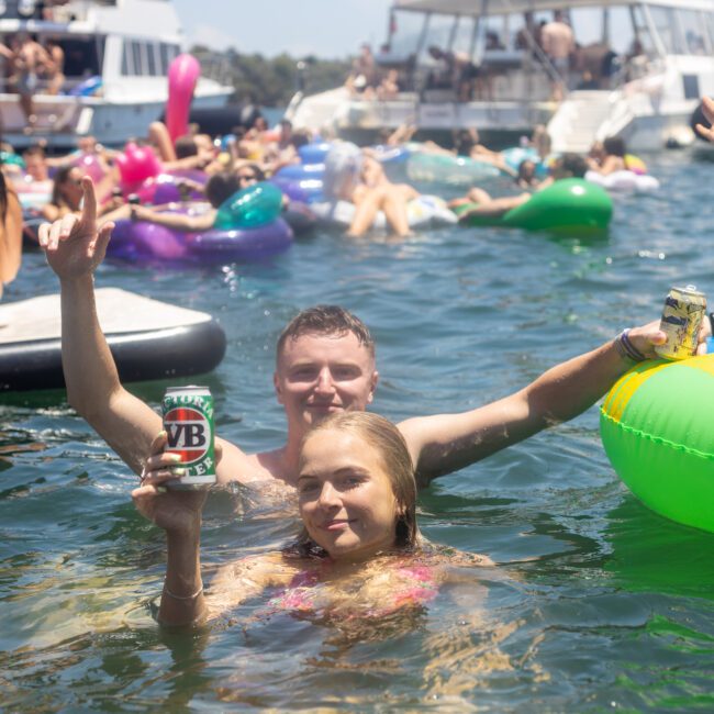 A man and woman in swimsuits stand waist-deep in clear water, each holding a can. They're surrounded by colorful inflatables and other people enjoying a sunny day on the water, with boats visible in the background.