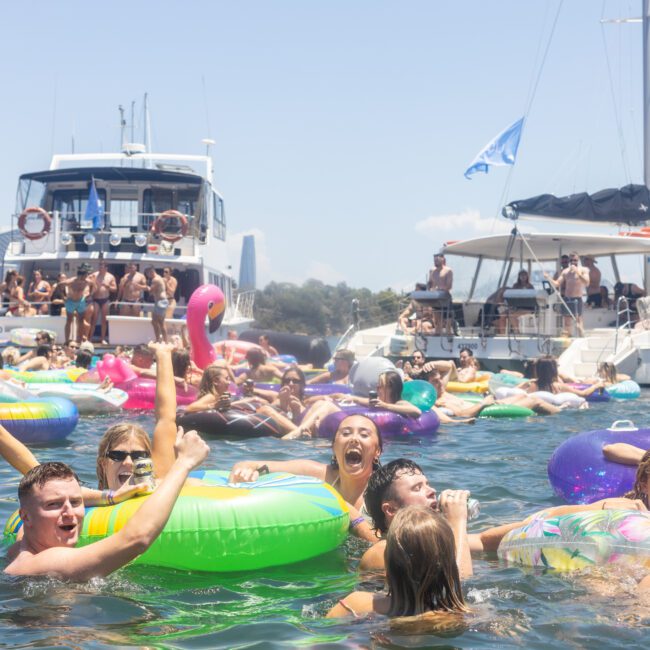 A group of people enjoying a sunny day on the water, floating on colorful inflatable tubes near two boats. The city skyline is visible in the background. The scene is lively and festive, with many smiling and having fun.