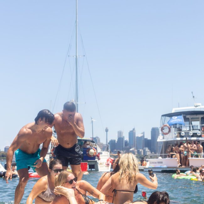 A group of people in swimwear are enjoying a sunny day on a floating mat on the water. The city skyline and boats are visible in the background, and the atmosphere is lively and festive.