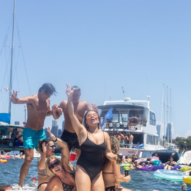 A group of people enjoying a sunny day on the water. They are on an inflatable platform, laughing and dancing, surrounded by boats and other floatation devices. The scene is lively and festive, set against a clear blue sky.