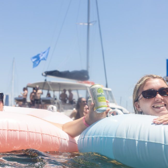 Two people relax on inflatable tubes in the water, each holding a drink. Boats and people are visible in the background. It's a sunny day, and both are wearing sunglasses, enjoying a fun time outdoors.