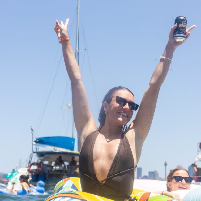 A woman in sunglasses and a swimsuit raises her arms in celebration while holding a can, floating on water with boats and blue sky in the background.