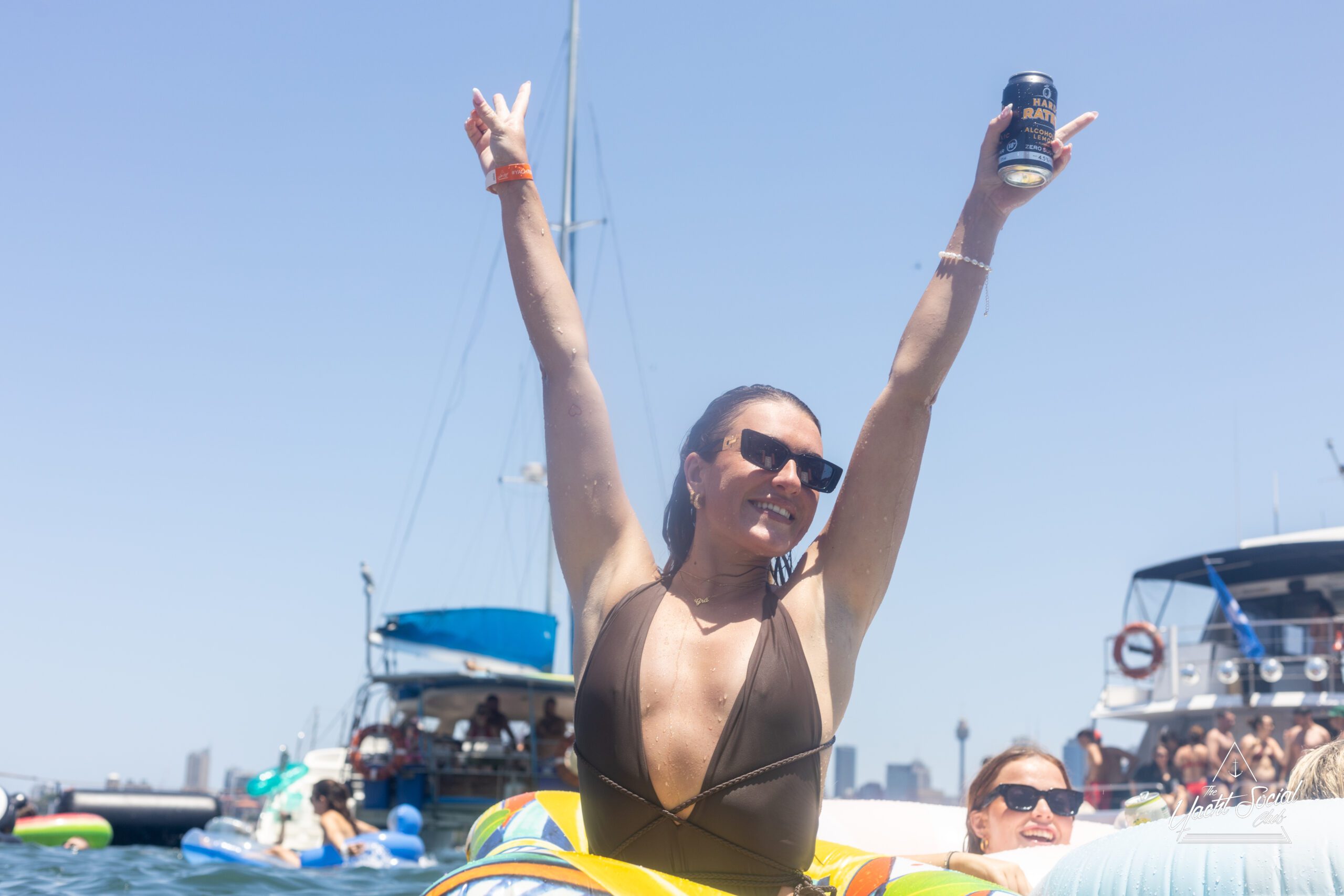 A woman in sunglasses and a swimsuit raises her arms in celebration while holding a can, floating on water with boats and blue sky in the background.