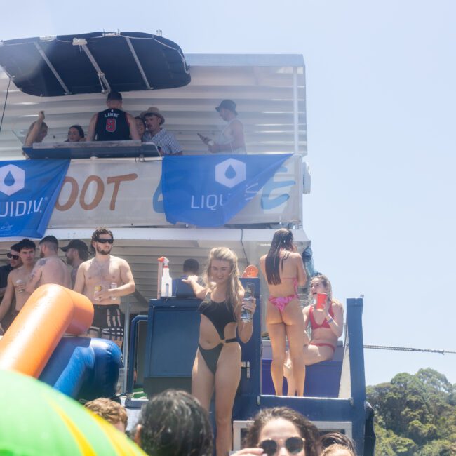 A group of people enjoying a sunny day on a houseboat, some in swimsuits. There are banners with the word "Liquid." A woman is posing with a drink, and inflatable toys are in the foreground. The sky is clear and blue.