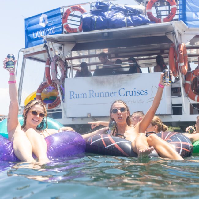 People relaxing on inflatable floats in the water near a boat with a "Rum Runner Cruises" sign. Two women in swimsuits hold drinks and smile, enjoying a sunny day. Life preservers and a lively atmosphere are visible.