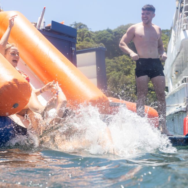 People enjoying a sunny day on the water. A person slides down an inflatable slide into the water, splashing around, while others watch from the boat. Trees and a clear sky are visible in the background.