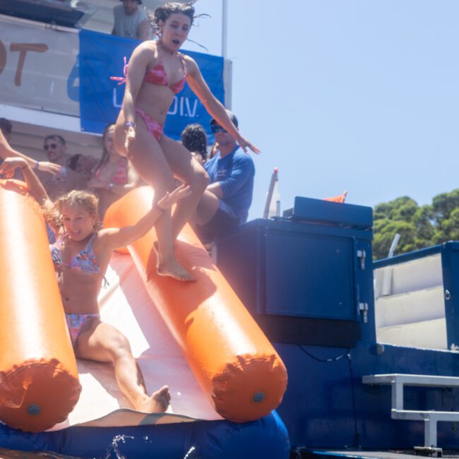 Two people are sliding down an inflatable slide from a boat into the water. They are in swimsuits, and the scene is filled with bright sunlight and a clear blue sky. Other people are watching from the boat deck.