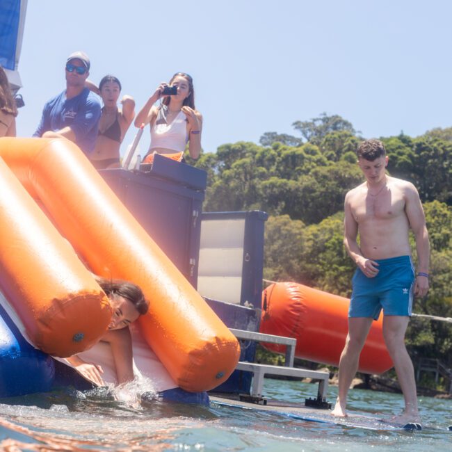 A group of people on a boat enjoying a sunny day. One person slides off an orange and blue water slide into the clear water below. Others watch and take photos. Lush greenery is visible in the background.