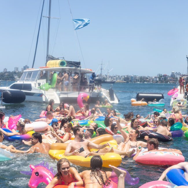 A lively gathering of people in colorful inflatable rings and floats in the water near a sailboat. The scene is festive and crowded, with a blue sky and city skyline in the background.