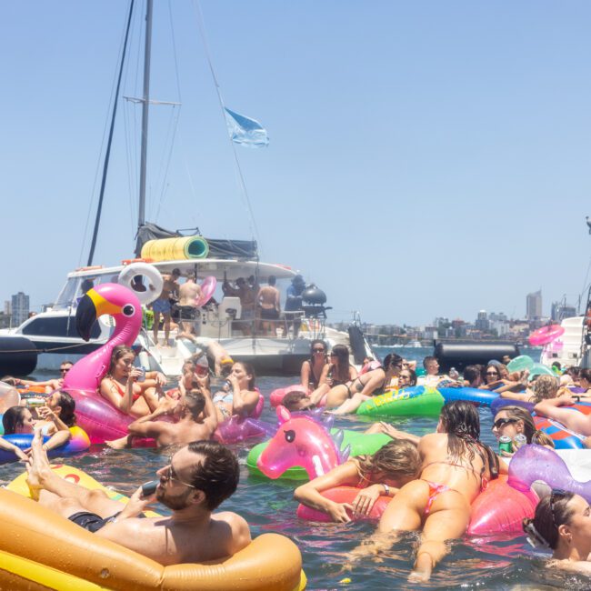 A crowd of people enjoying a sunny day in the water on a variety of colorful inflatable rafts, including flamingos and unicorns, with boats and a city skyline visible in the background.