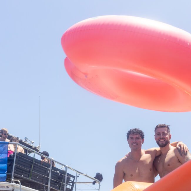 Two people enjoying a sunny day on a boat with a large orange inflatable ring overhead. They are smiling, with others seen in the background also on the boat. Clear blue sky completes the scene.