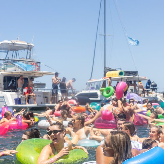 A lively scene at a boat party with many people enjoying the water on inflatable floats of various colors and shapes. Boats are anchored nearby, and the sky is clear and sunny.