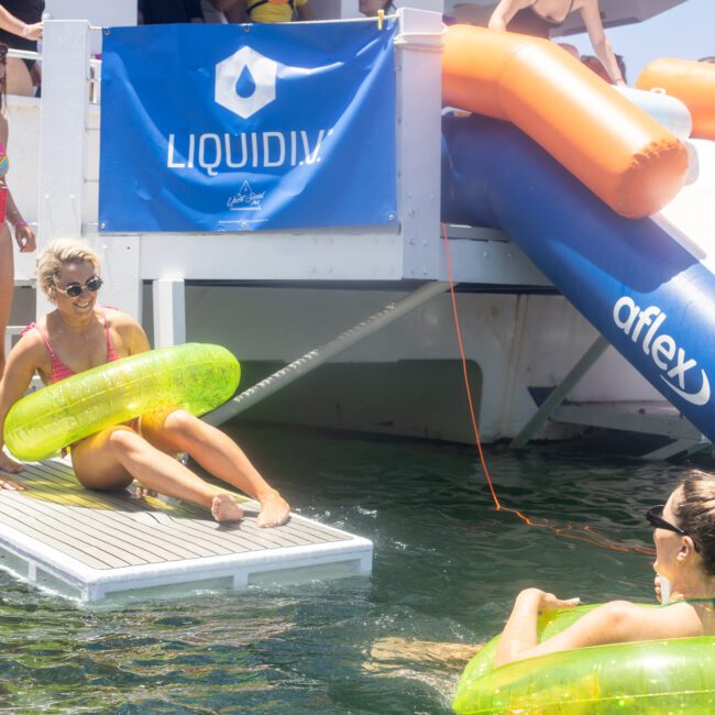 Three people enjoy a sunny day on a floating platform. One is about to enter the water using a small slide, while another stands beside her. A third person floats in the water, all wearing swimsuits and engaged in a lively atmosphere.