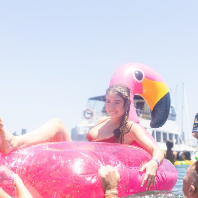 A woman relaxes on a pink flamingo float in the water, surrounded by boats under a clear sky. People enjoy the sunny day, with one holding a can. The scene captures a cheerful, summer vibe at a lively gathering.