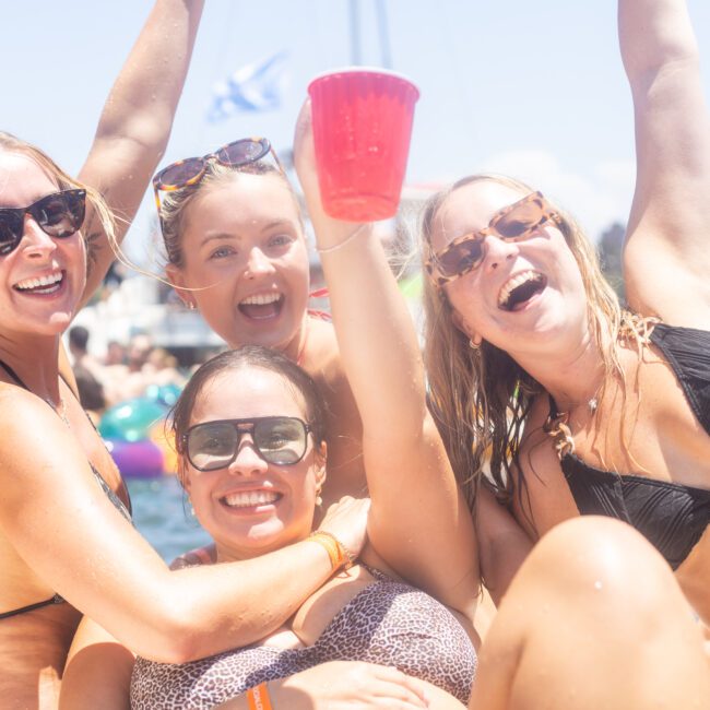 Four women in swimsuits enjoy a sunny day on a boat, smiling and raising a red cup. The background features a blue sky, sea, and part of a boat with flags. Everyone appears to be celebrating or having fun.