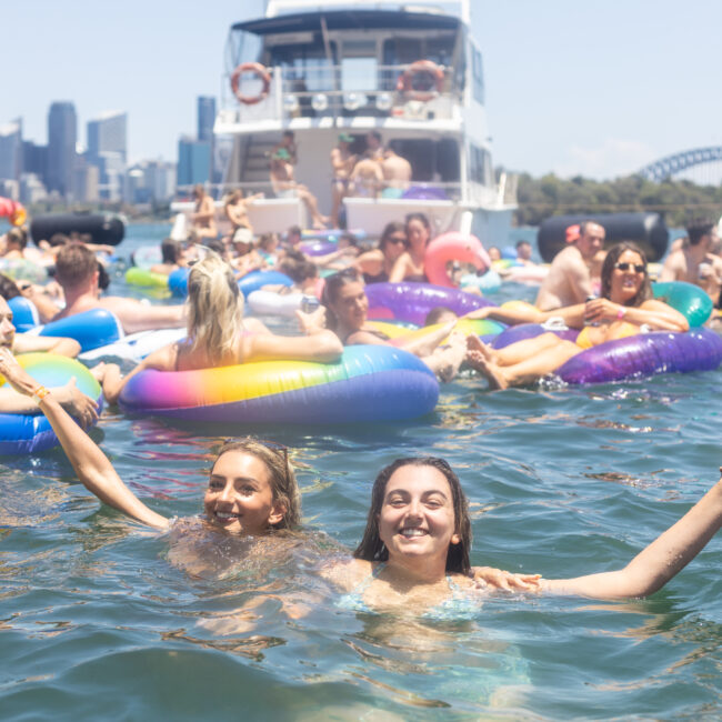 A group of people enjoying a sunny day in the water, surrounded by colorful inflatable pool floats. In the background are boats and the skyline of a city with a bridge. Two smiling individuals pose for the camera in the foreground.