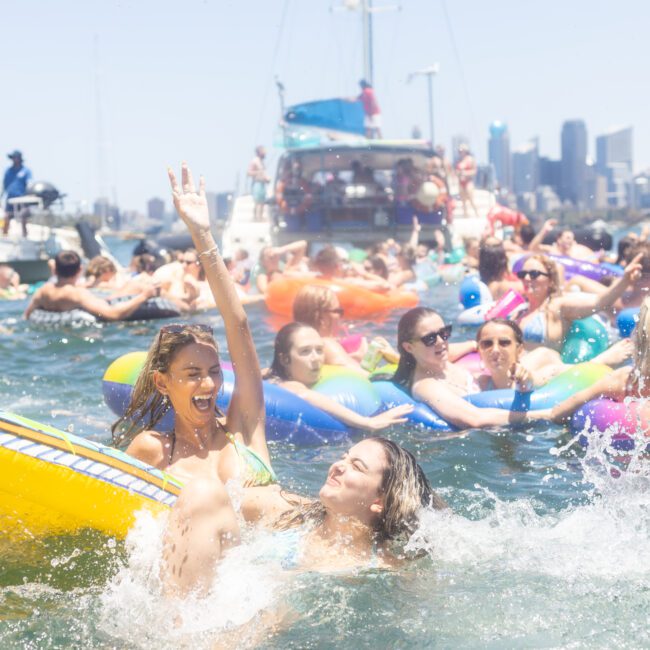 A lively crowd enjoys a sunny day on the water near a city skyline. People relax on colorful inflatable rafts and boats, laughing and splashing in the vibrant scene.