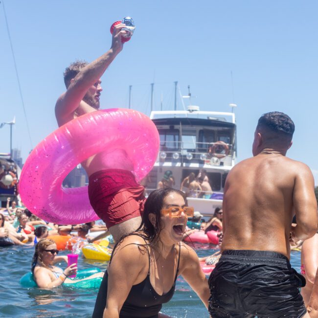 People are having fun at a lively pool party. A man with a pink inflatable ring splashes into the water while others float around. A boat is visible in the background under a clear blue sky.