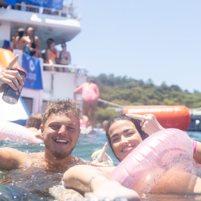 A smiling man holding a drink and a woman lounging on a pink float enjoy their time in the water near a boat. People can be seen on the boat in the background, with inflatable slides leading into the sea. It's a sunny day.